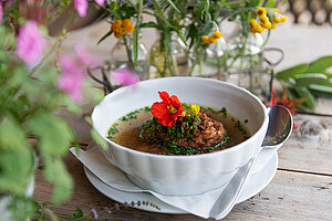 Genuss-Tipp der Woche Eine köstliche Schüssel Kaspressknödelsuppe, garniert mit frischen Kräutern und bunten essbaren Blumen, steht auf einem rustikalen Holztisch, umgeben von zarten Blumenarrangements, die eine einladende Atmosphäre schaffen.
