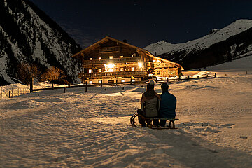 Ein Pärchen sitzt bei Nacht auf einem Schlitten im Schnee, mit Blick auf ein uriges, beleuchtetes Bauernhaus